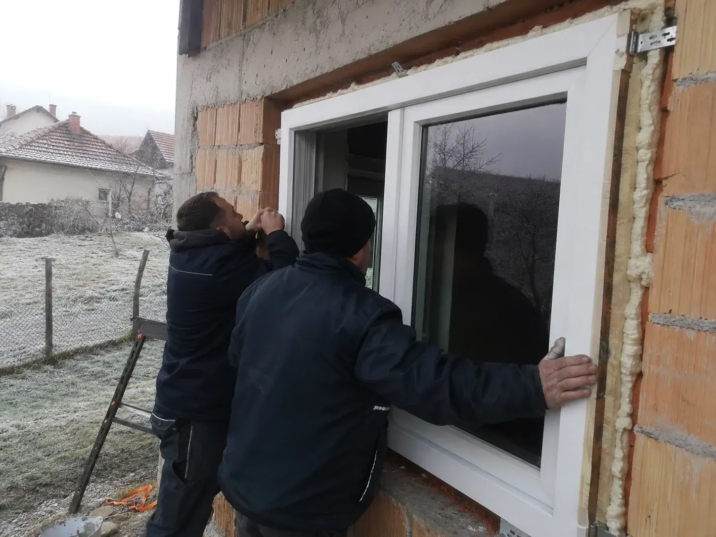 Dva montera u zimskim radnim jaknama ugrađuju PVC prozor, mraz na zemlji napolju.