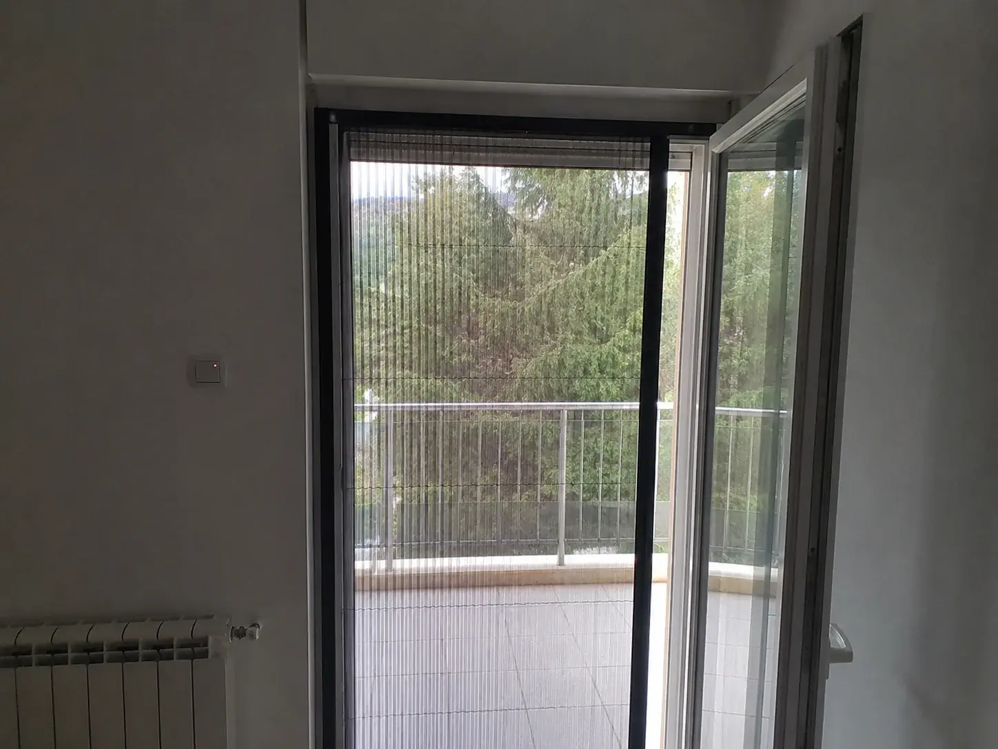 Plise komarnik delimično razvučen preko balkonskih vrata stana, pogled iz sobe ka terasi