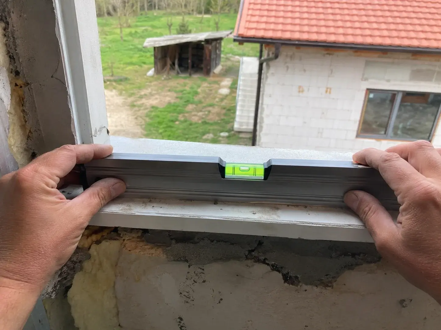 Ruke drže libelu horizontalno na donjem rubu otvora prozora, mehurić blago pomeren.