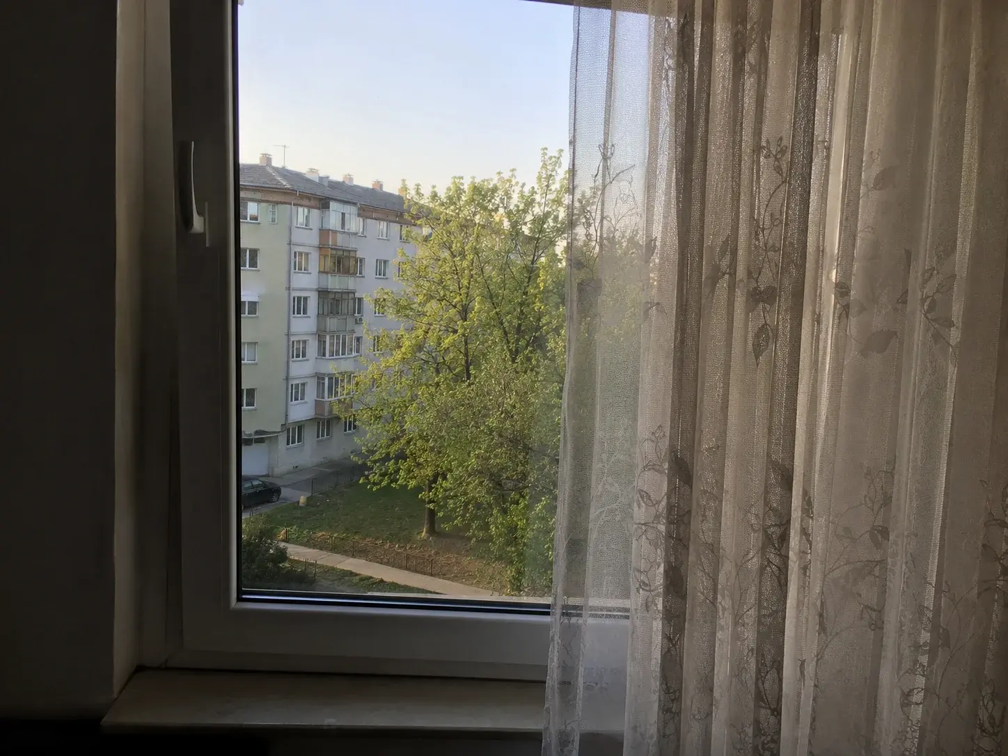 Mirna spavaća soba sa prozorom ka tihoj rezidentnoj ulici, jutarnje svetlo, providna zavesa.